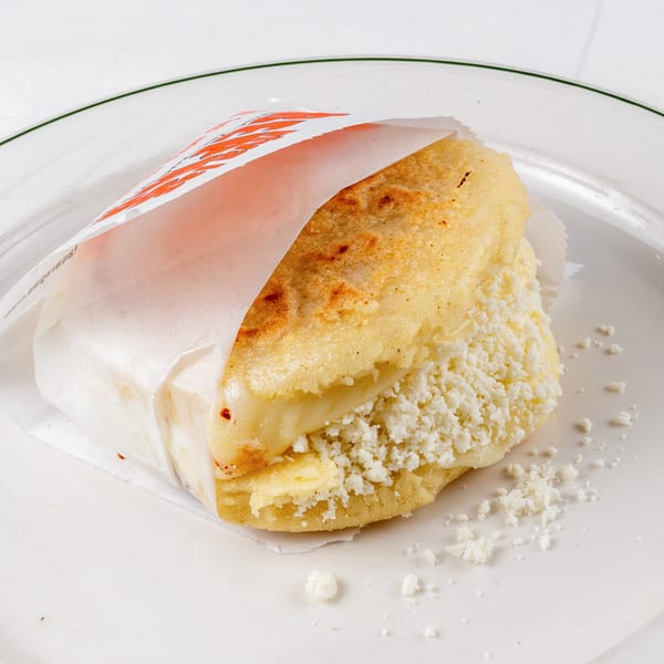A Viva Las Arepas cheese arepa, wrapped in paper, sits on a white plate.