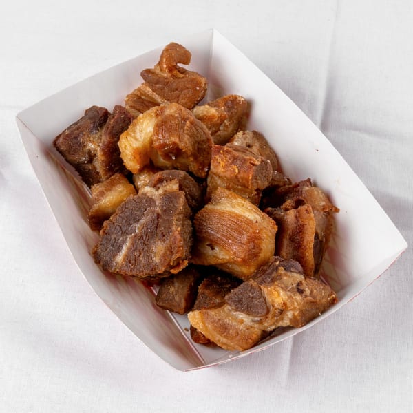 Crispy, golden-brown fried pork chunks, chicharrones, in a white paper tray on a white tablecloth.
